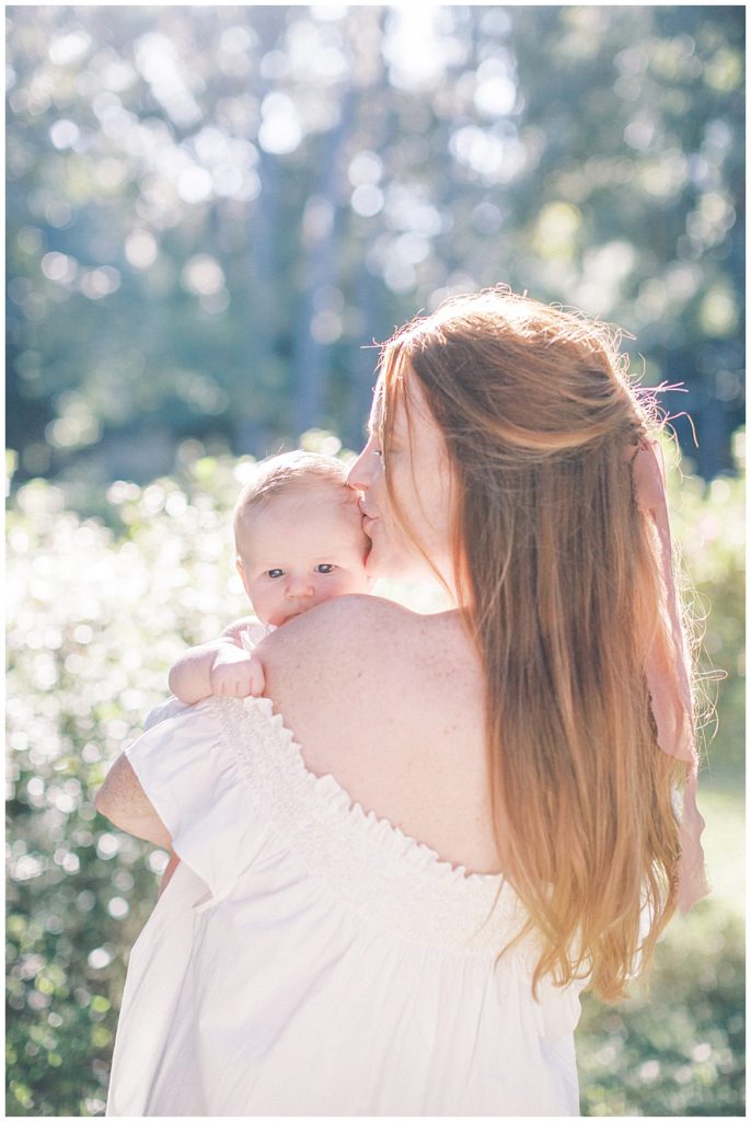 A Red Haired Mother Holds Her Newborn Baby Up To Her Shoulder And Turns Her Head To Kiss The Side Of His Head During Their Outdoor Newborn Session