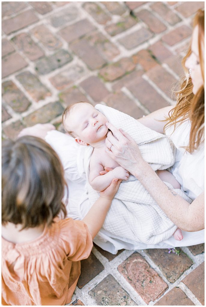 A Mother Sits On Brick Steps Holding Her Newborn Next To Her Young Daughter While They Both Gently Touch The Baby's Face