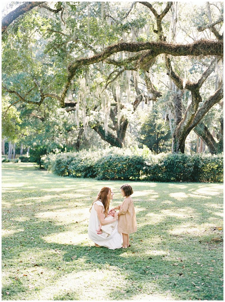 A Mother Knees In A Field Holding Her Baby While Her Daughter Comes Over To Caress The Baby