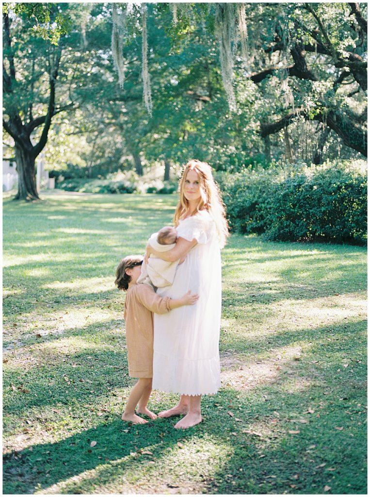 A Red Haired Mother Stands In A Green Park Holding Her Newborn Baby Boy While Her Daughter Stands In Front Of Her Hugging Her