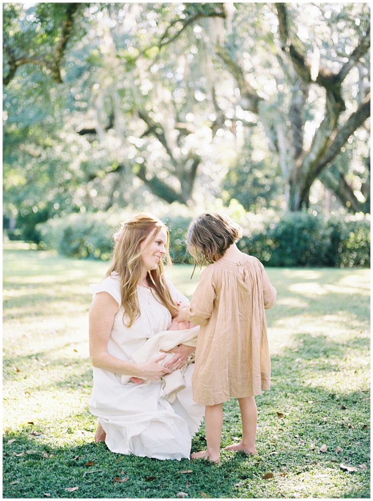 A Mother Kneels Down In A Green Park Holding Her Newborn Up To Her Young Daughter Who Looks Lovingly At The New Baby