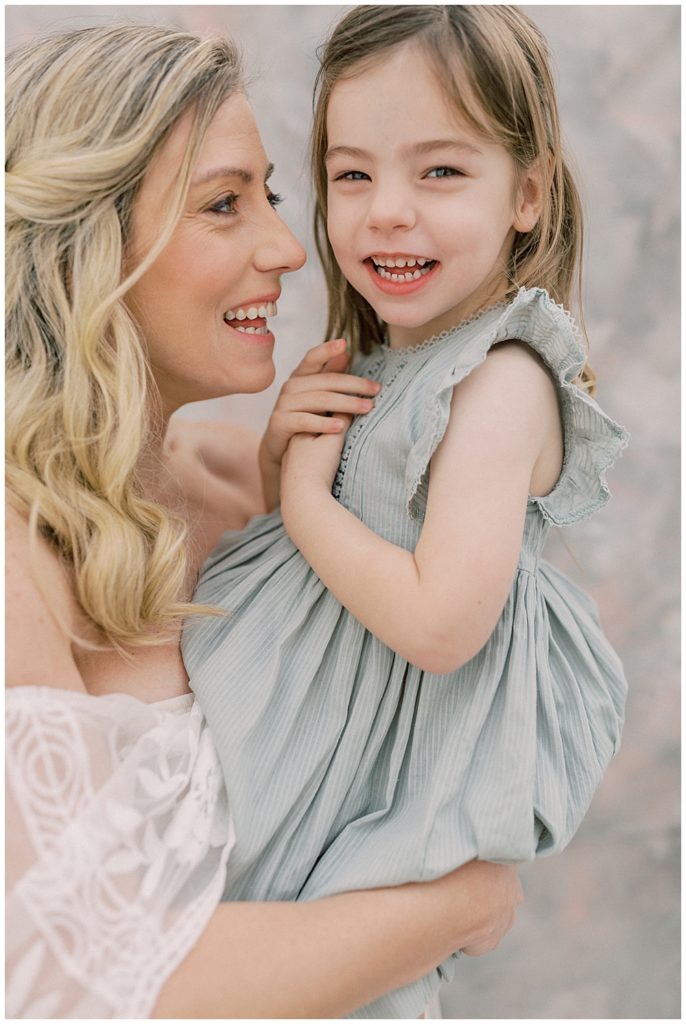 A Little Girl In A Green Dress Sits On Her Mother's Lap While Her Mother Smiles At Her