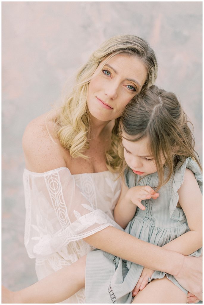 A Blonde Mother Holds Her Little Girl Up Against Her And Smiles Softly At The Camera