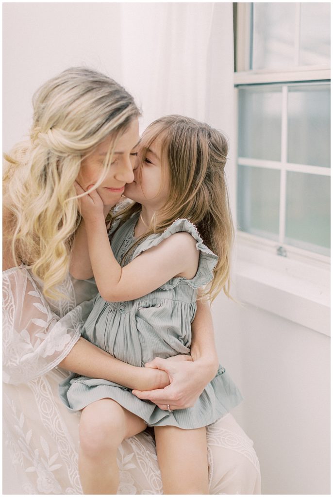 A Little Girl In A Green Dress Whispers In Her Mother's Ear While Sitting In Her Lap Near A Window