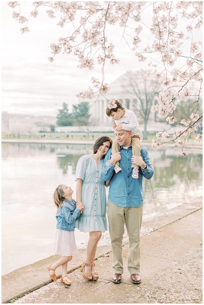 Mother Rests Her Head On Her Husband's Shoulders While Her Daughter Stands Next To Her And Son Sits On Her Husband's Shoulders. They Are Standing By The Tidal Basin In Dc