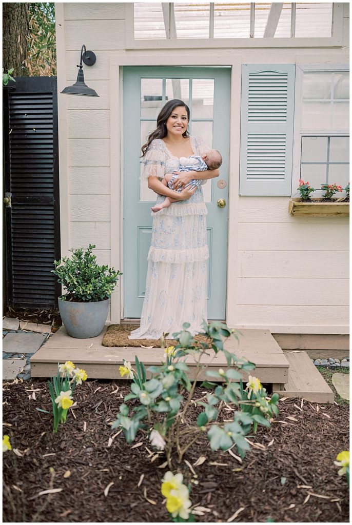 A Mother Holds Her Infant Son While Wearing A Blue Needle And Thread Gown Standing In Front Of A Blue Door