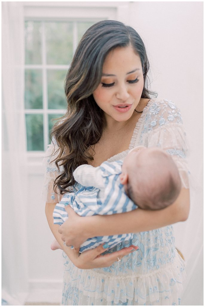 A Mother Wearing A Blue Needle And Thread Dress Looks Down And Comforts Her Infant Son During Her Motherhood Studio Session