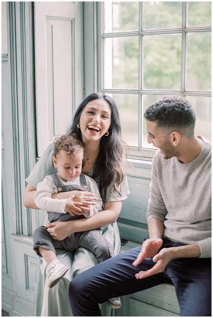 Mother Laughs While Holding Her Toddler Son With Her Husband In The Window At Salubria