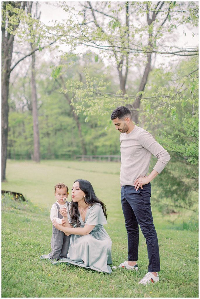 A Mother Blows A Dandelion With Her Toddler Son While The Dad Stands And Watches