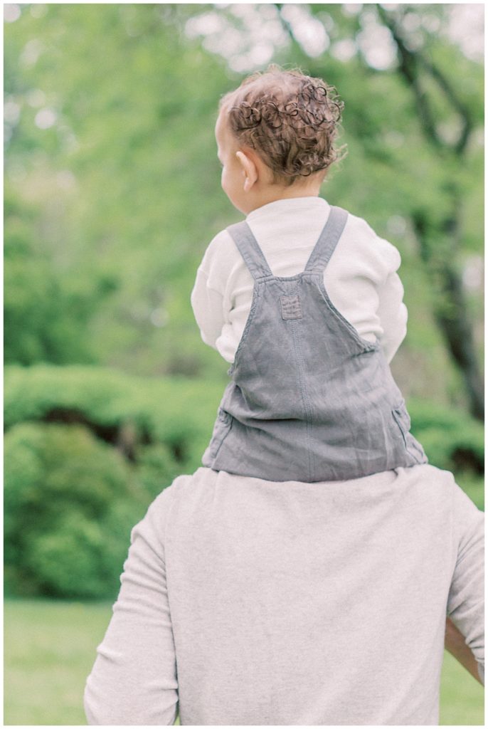 Toddler boy in overalls rides on his dad's shoulders