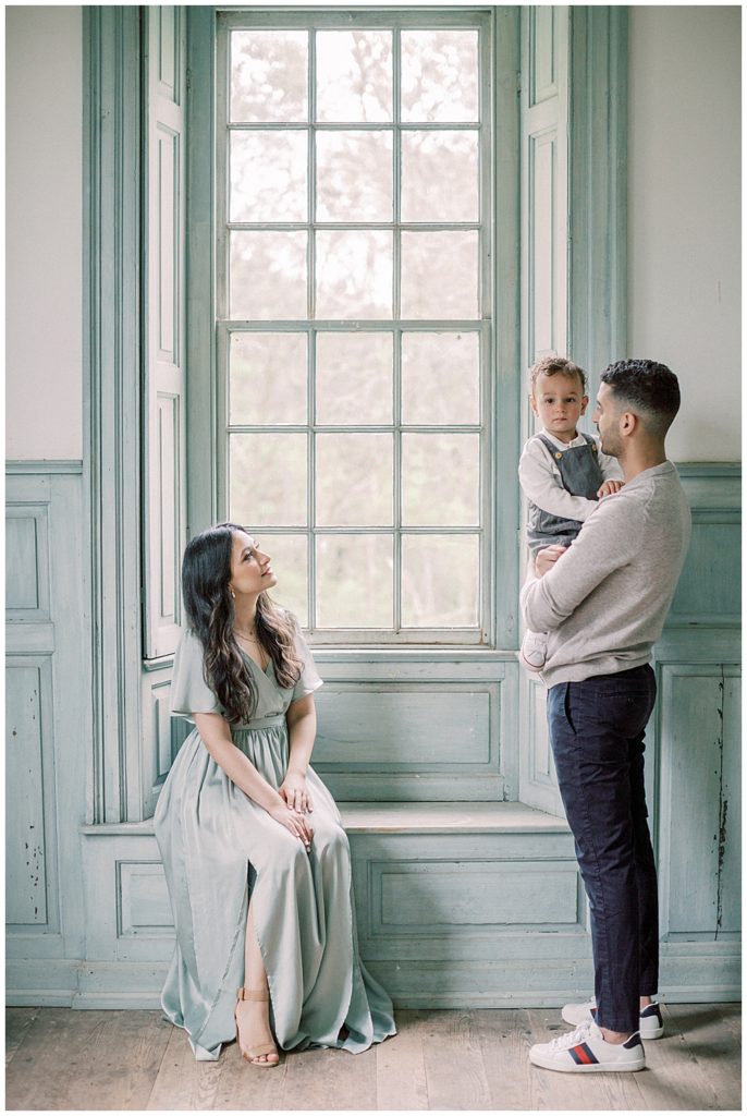 A Mother In A Blue Dress Sits In The Window At Salubria While Looking Up At Her Husband Who Is Holding Their Toddler Son