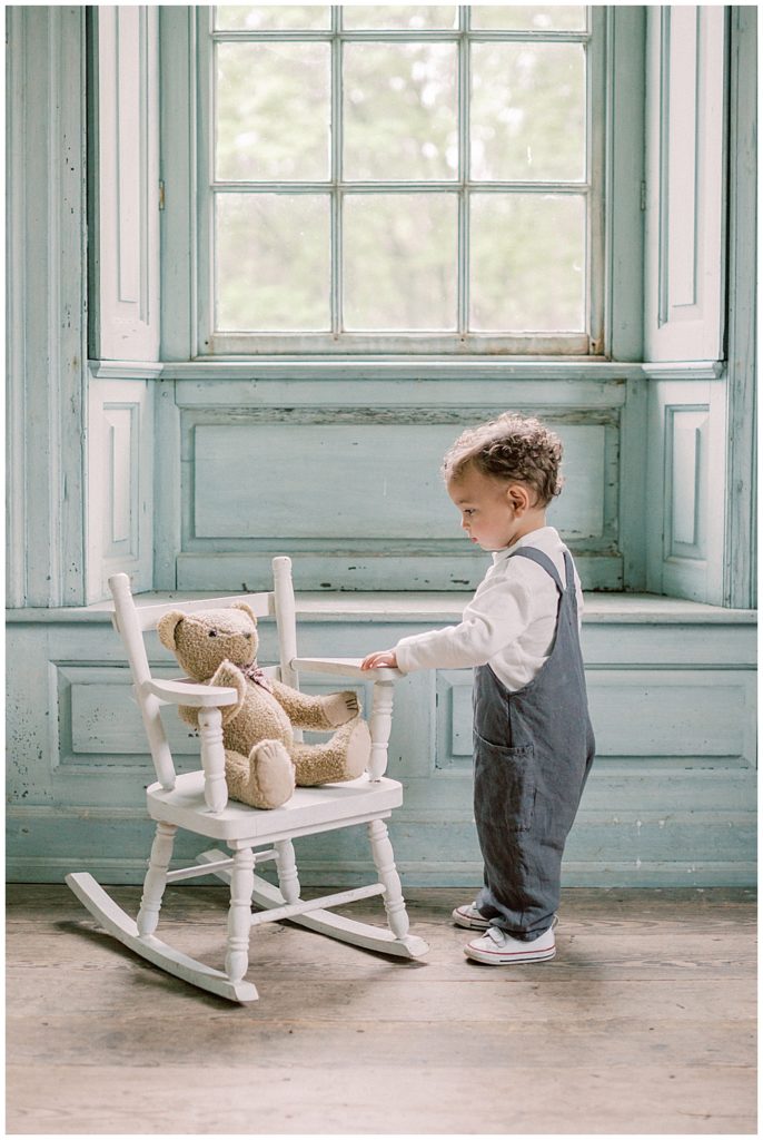 Toddler Boy Stands In Front Of A Small White Rocking Chair With A Teddy Bear On It In Front Of A Large Window At Salubria