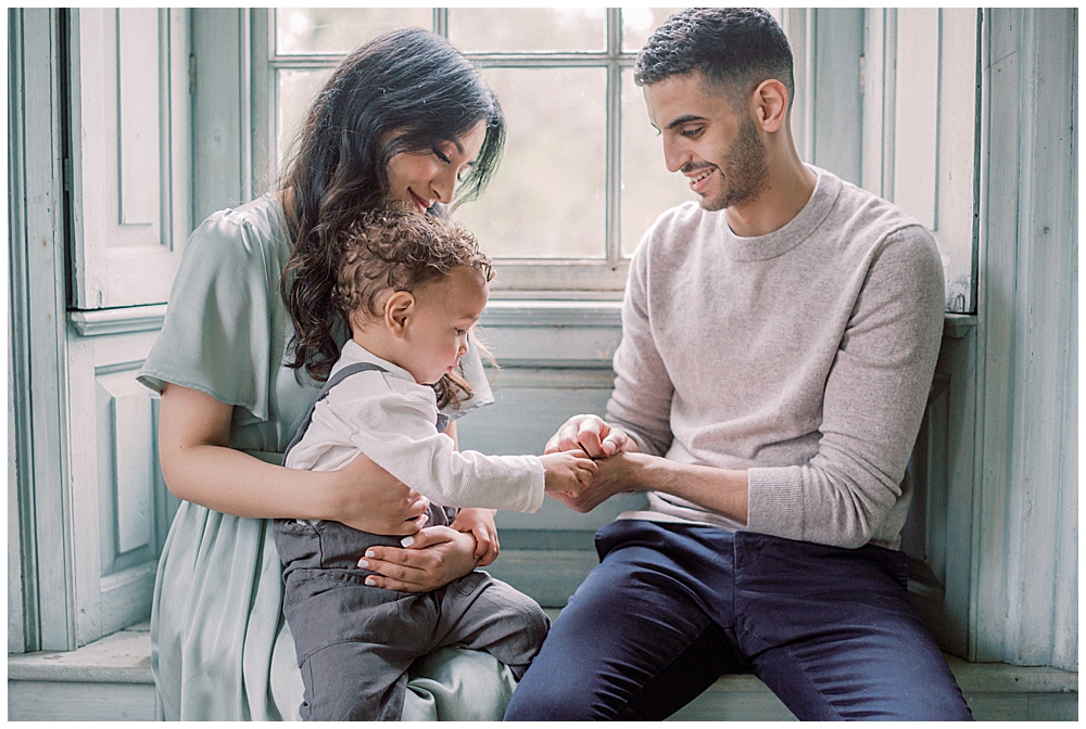 Mother and father sit in front of a window with their toddler son on their lap in Salubria, Virginia