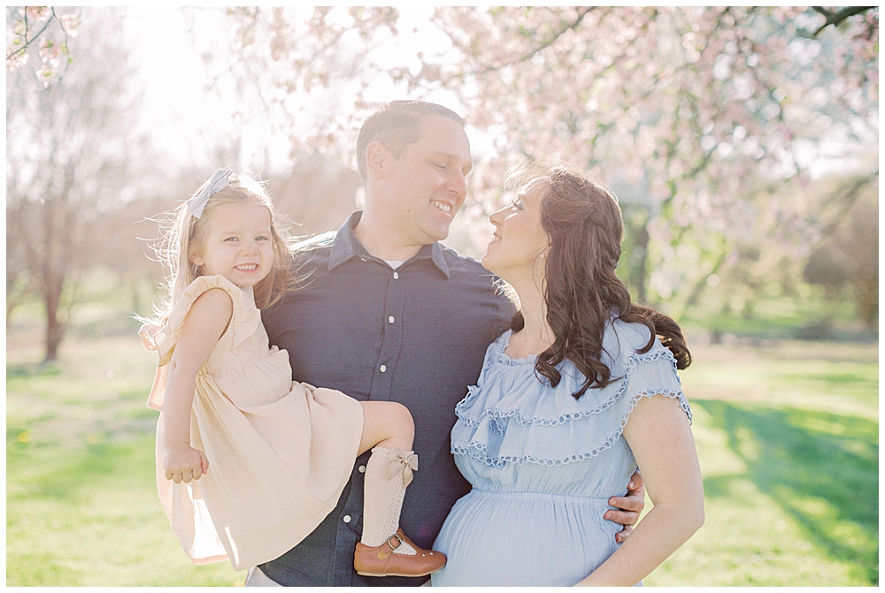 A Mother And Father Hold Their Toddler Daughter And Look At Each Other Smiling