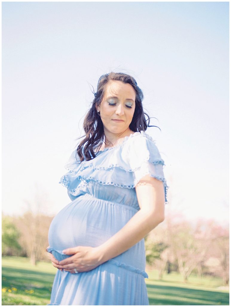 A Pregnant Mother Wearing A Blue Dress Stands With Her Eyes Closed, Holding Her Belly.