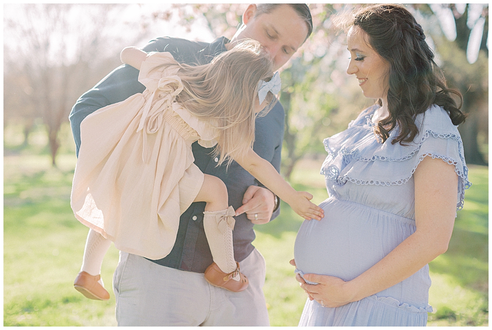 A Little Girl, Held By Her Father, Leans Down And Touches Her Mom's Pregnant Belly