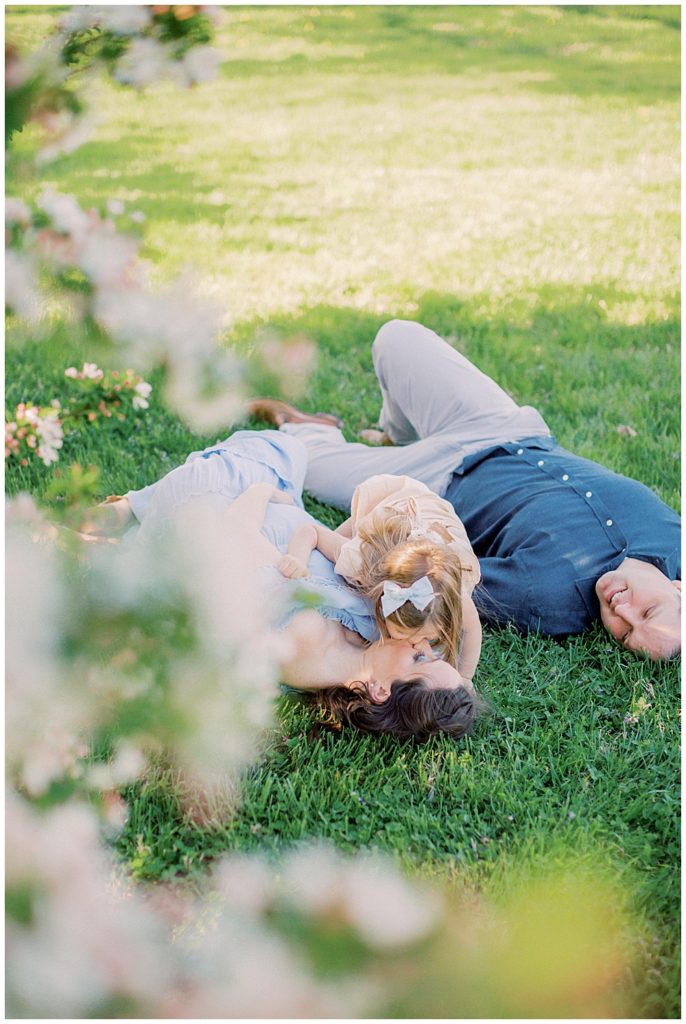 A Little Girl Leans Down And Kisses Her Mother While Lying In The Grass 