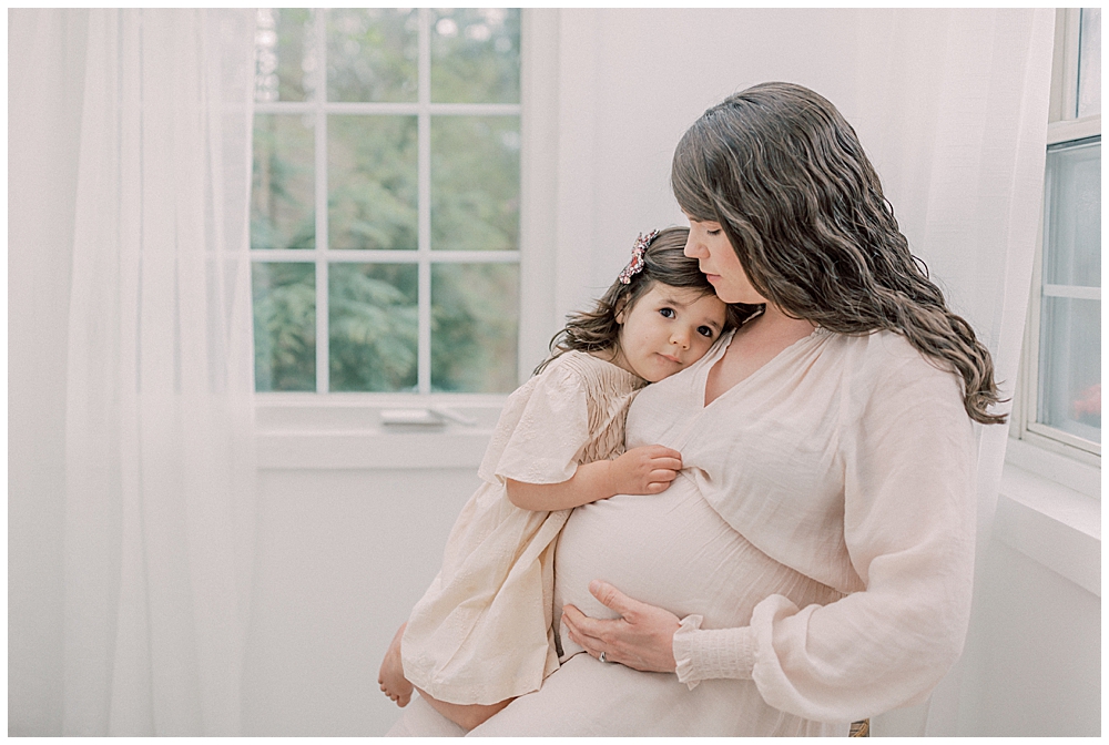 An Expecting Mother Sits On A Stool While Holding Her Toddler Daughter Who Rests Her Head On Her Mom.