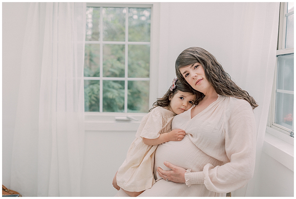 Little Girl Sits With Her Head Resting On Her Mother During Their Studio Maternity Session