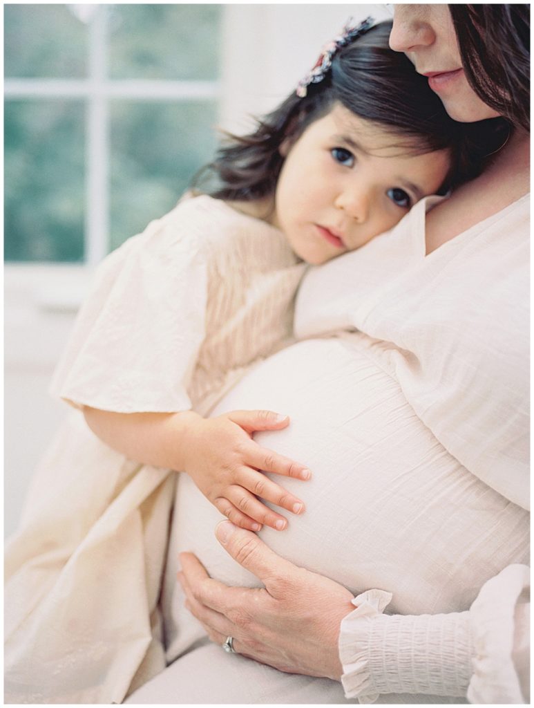 Little Girl Lays Her Head On Her Mother's Chest And Rests Her Hand On Her Mother's Pregnant Belly