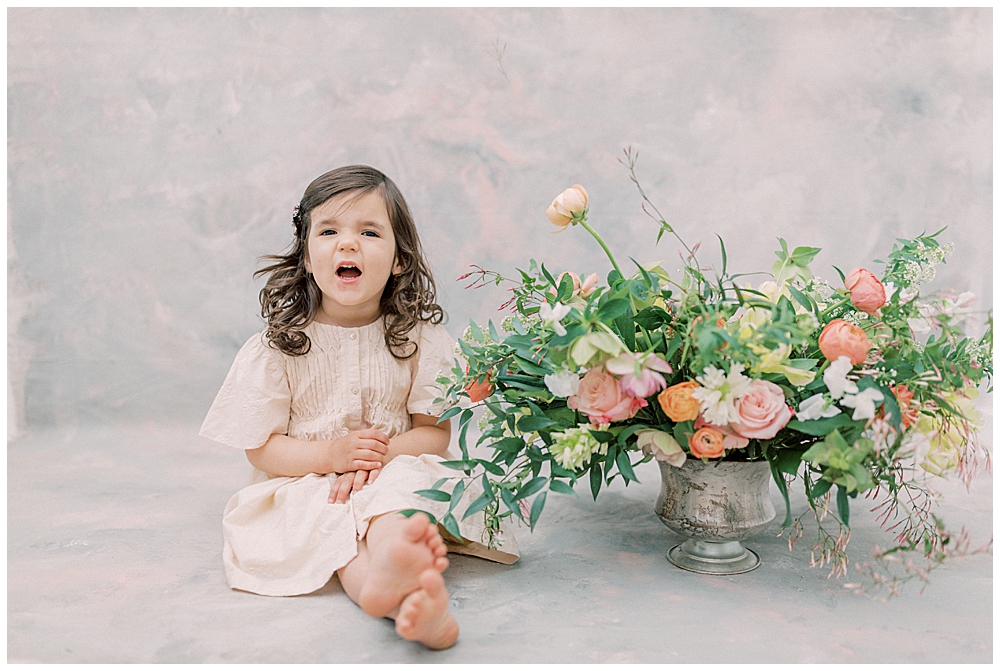 Little Girl Sits Next To Floral Display And Smiles In A Photography Studio