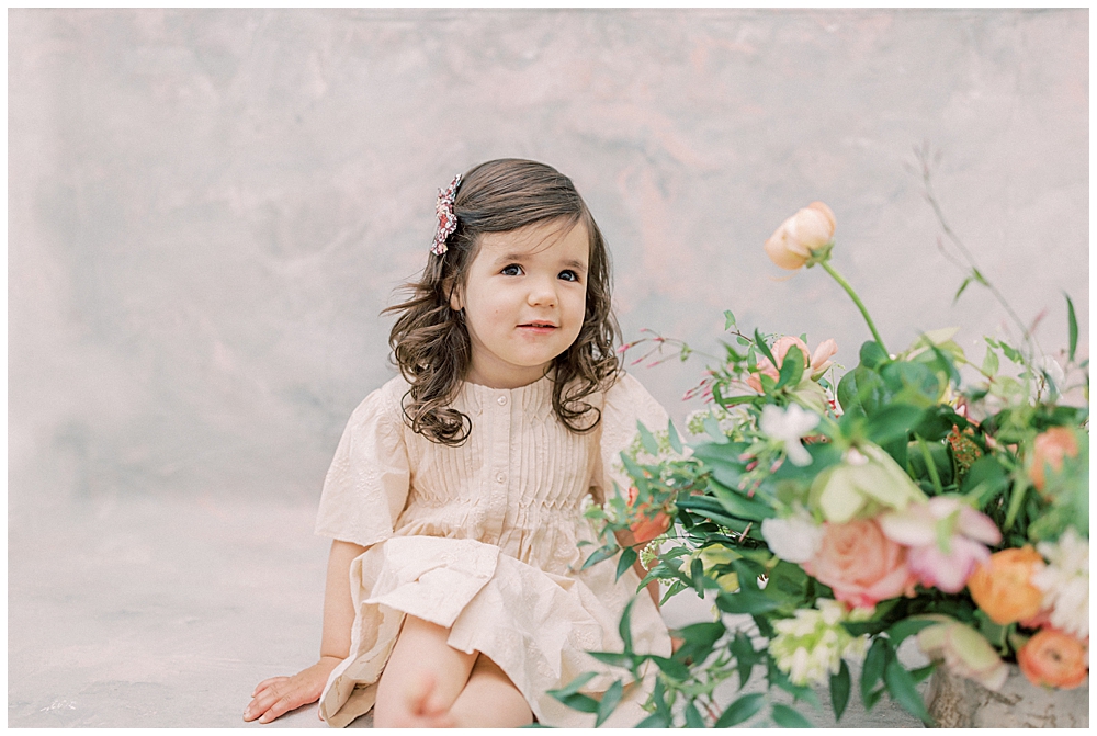 Little Girl Sits Next To Flowers In Vase And Smiles