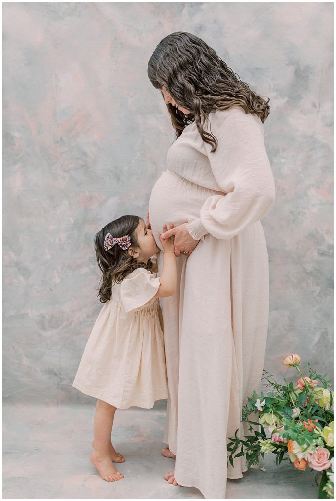 A Toddler Girl Kisses Her Mother's Belly During Their Maternity Studio Session With Daughter And Mother 