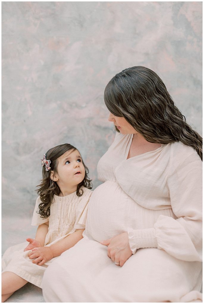 Toddler Girl Sits With Her Mother And Looks Up At Her During Maternity Studio Session With Daughter And Mother