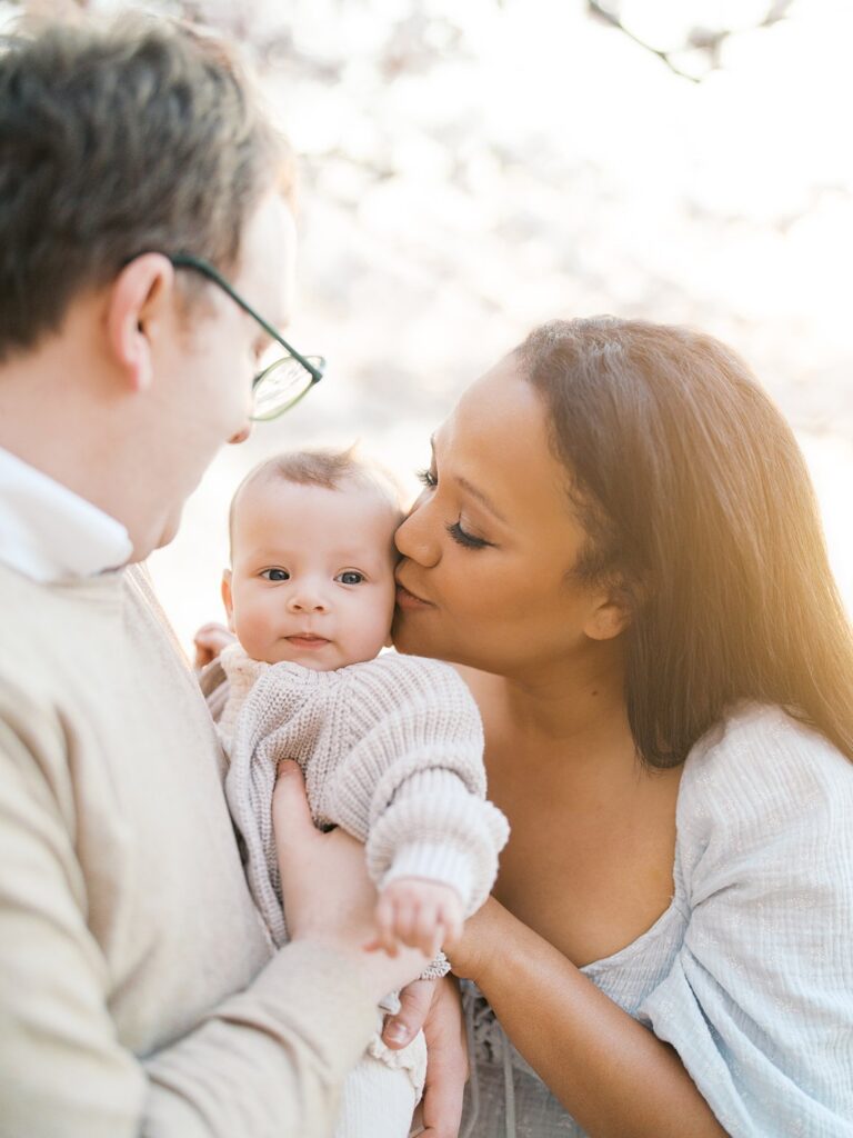 A Close-Up Of A Mother Kissing Her Baby's Cheek While Her Partner Looks On, Warm Golden Light And Tidal Basin Cherry Blossoms Glowing Softly Behind Them.