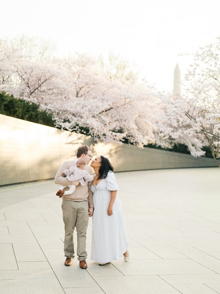 A Family Of Three Stands Together On The Plaza With The Washington Monument In The Distance, Surrounded By Tidal Basin Cherry Blossoms In Full Bloom At Golden Hour.
