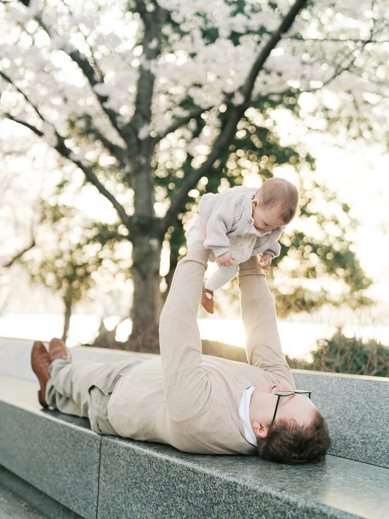 A Father Lies On His Back Along A Stone Ledge Near The Tidal Basin Cherry Blossoms, Lifting His Giggling Baby Into The Air Above Him With Trees Glowing Golden Behind Them.