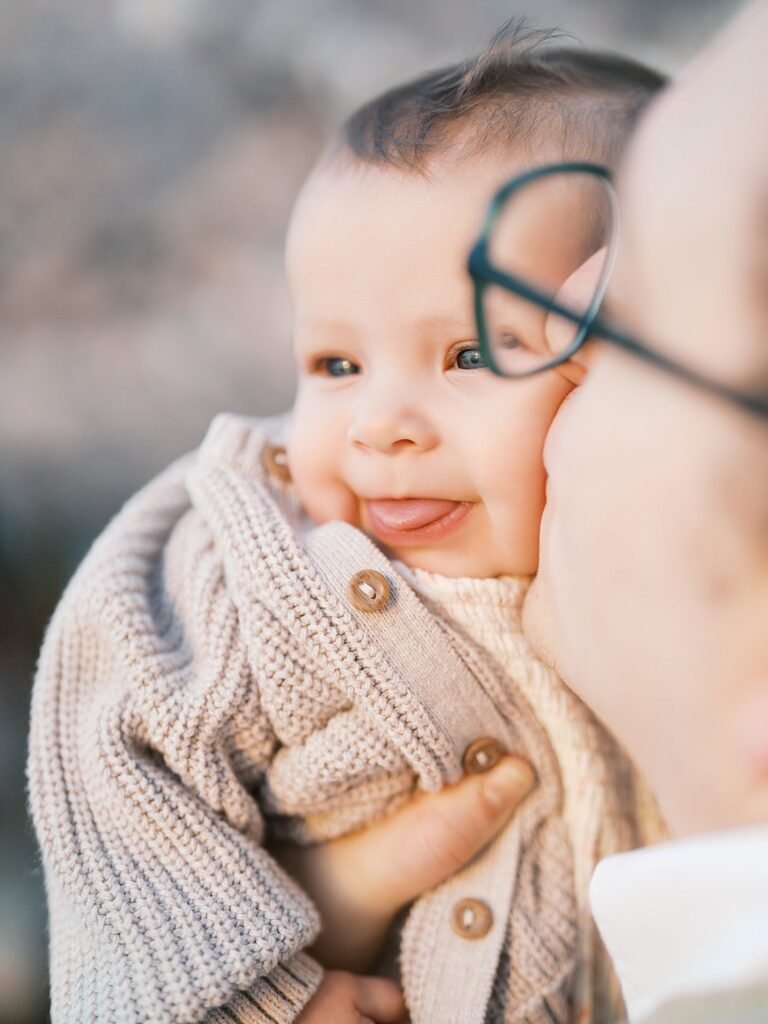 A Close-Up Of A Smiling Baby Snuggled Against A Parent's Cheek, Wearing A Cozy Knit Cardigan With Cherry Blossoms Softly Blurred In The Background.