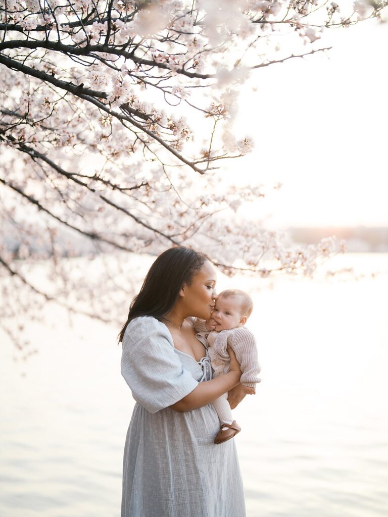 A Mother Kisses Her Baby's Cheek While Standing Alone Beneath Tidal Basin Cherry Blossoms, The Pale Pink Blooms And Shimmering Water Creating An Ethereal Backdrop Behind Her.