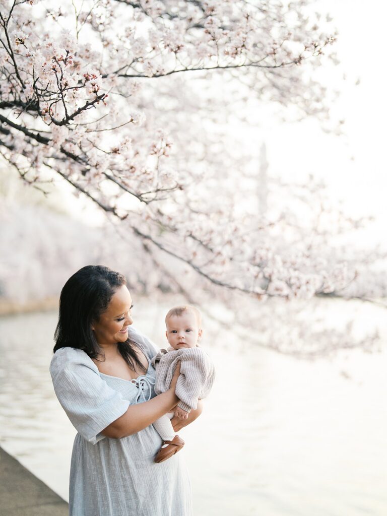 A Mother Smiles Down At Her Baby Boy While Standing Along The Water's Edge, Tidal Basin Cherry Blossoms Glowing Pink And White In The Dreamy Background.