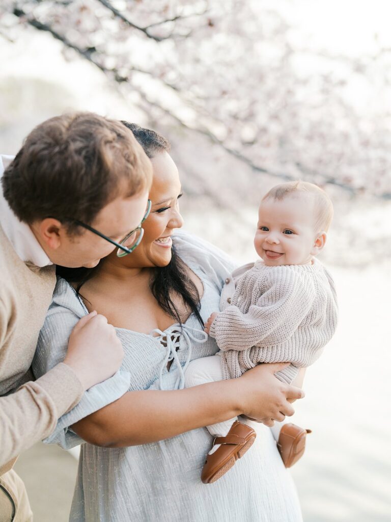 A Father Wraps His Arms Around His Partner From Behind As She Holds Their Smiling Baby, The Three Of Them Glowing In Warm Light Beneath The Tidal Basin Cherry Blossoms.