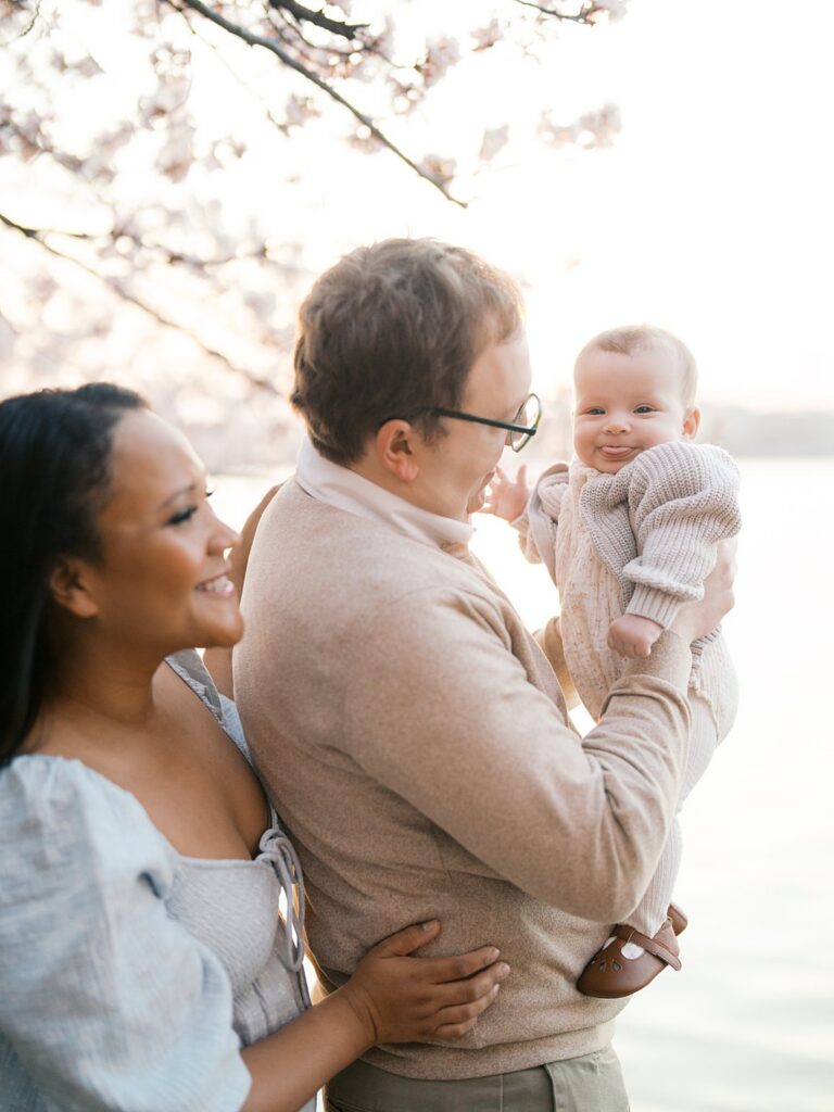 Two Parents Smile Up At Their Grinning Baby As Dad Holds Him Aloft, All Three Backlit By Golden Hour Glow And Soft Tidal Basin Cherry Blossoms Overhead.