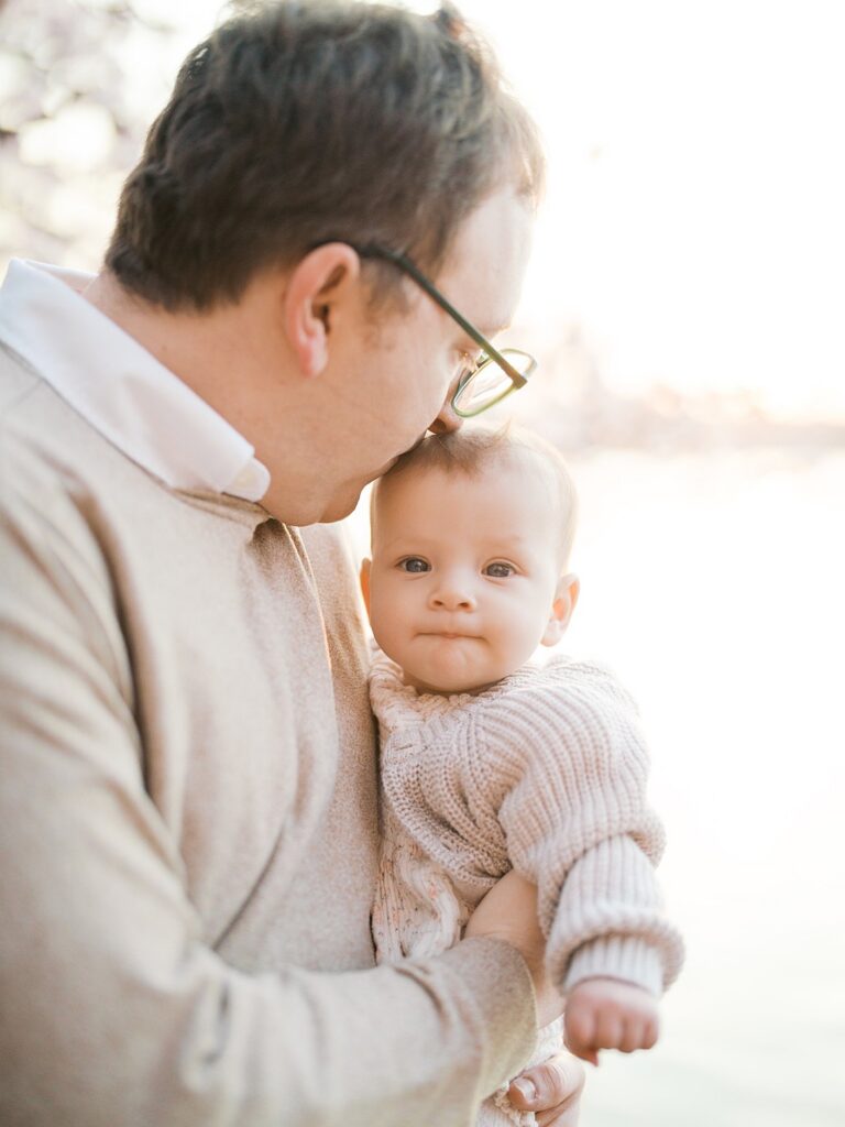 A Close-Up Of A Father Gazing Down At His Wide-Eyed Baby Nestled Against His Shoulder, Soft Cherry Blossom Light Wrapping Around Them Both.