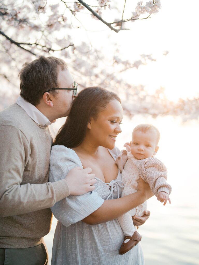 A Father Kisses His Partner's Temple While She Smiles Down At Their Baby, The Three Of Them Wrapped In Soft Light Beneath The Tidal Basin Cherry Blossoms.