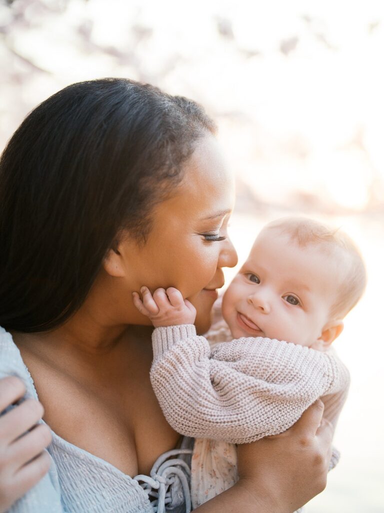 A Mother Gazes Lovingly At Her Alert Baby, Holding Him Close Cheek-To-Cheek In Soft Golden Light During A Tidal Basin Cherry Blossoms Family Session.