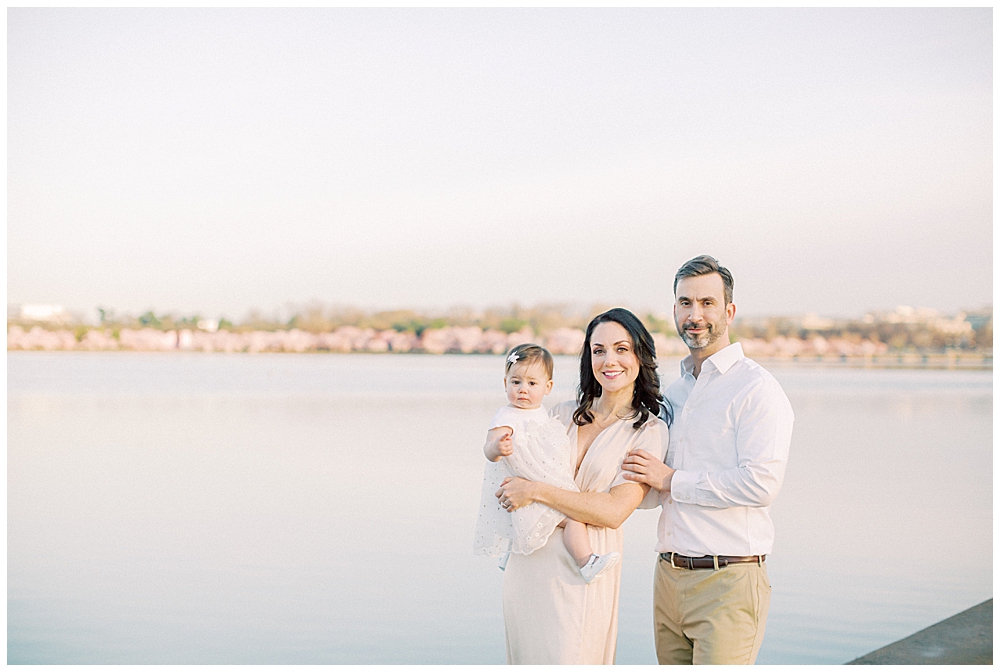 A Mother And Father Stand At The Tidal Basin With Their Daughter With The Cherry Blossoms Behind Them.