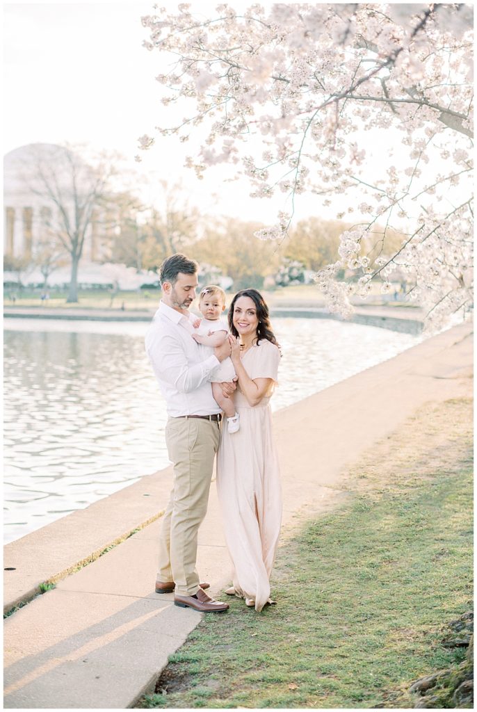 Mother Smiles At The Camera While Standing With Her Husband And One Year Old Daughter During Sunrise At The Tidal Basin Cherry Blossoms