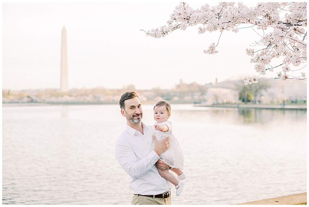 A Father Holds His Baby Daughter Next To A Cherry Blossom Tree Across From The Washington Monument In Dc