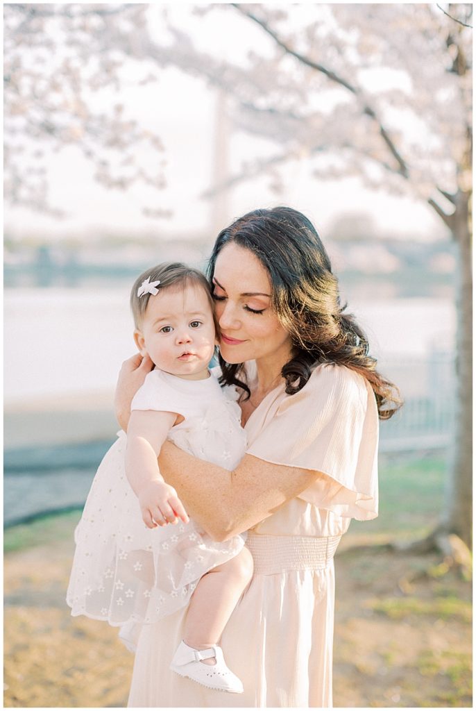 A Mother Brings Her Baby Daughter Up To Her Face And Snuggles Her Close While Standing Near Cherry Blossom Trees In Dc.