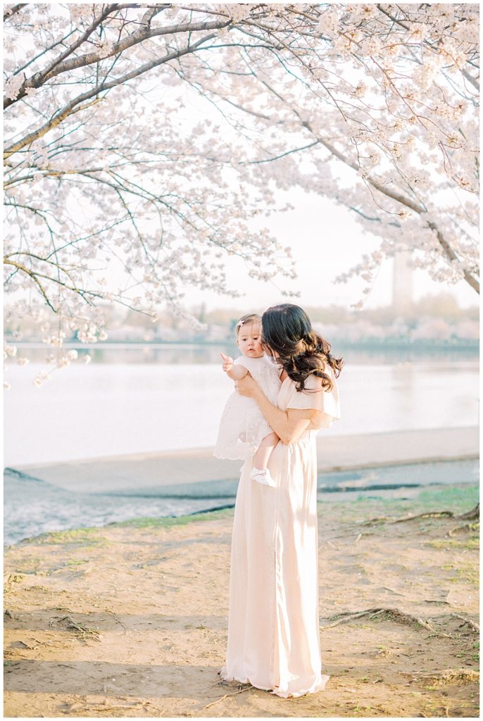 A Mother Wearing A Long Pink Gown Kisses Her Infant Daughter's Cheek While They Stand Below Cherry Blossom Trees Along The Tidal Basin In Washington Dc