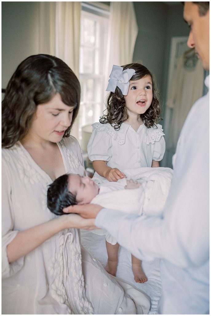 A Little Girl Smiles At Her Dad While Her Mom And Dad Hold Her Baby Sister On The Bed