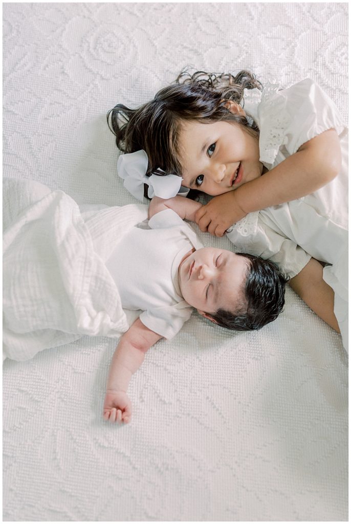 A Toddler Girl Lays On The Bed With Her Baby Sister, Holding Her Hand