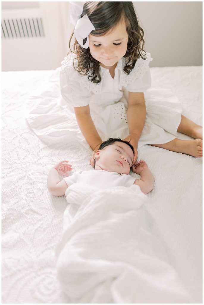 A Toddler Girl Cradles Her Baby Sister's Head On The Bed