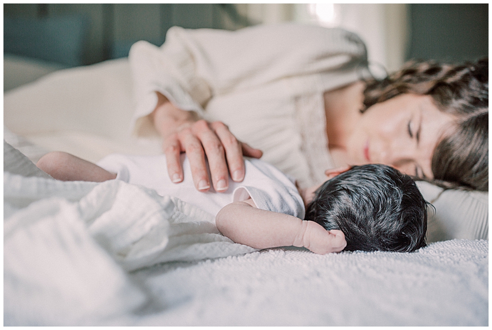 Newborn And Mother Lay On The Bed Together With The Mother's Hand Resting On Her Newborn's Chest.