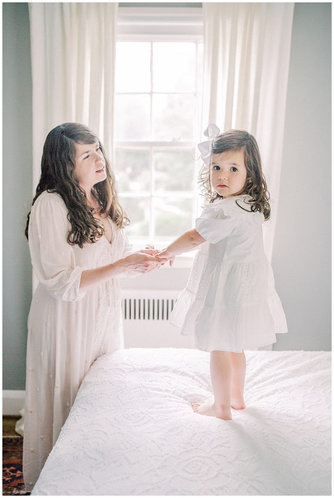 A Mother Holds Her Toddler Daughter's Hands While Her Toddler Stands On The Bed