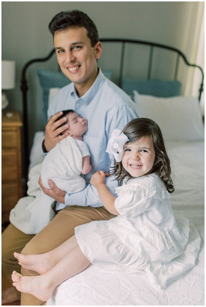 A Father Sits On The Bed With His Newborn And Toddler Daughters And Smiles